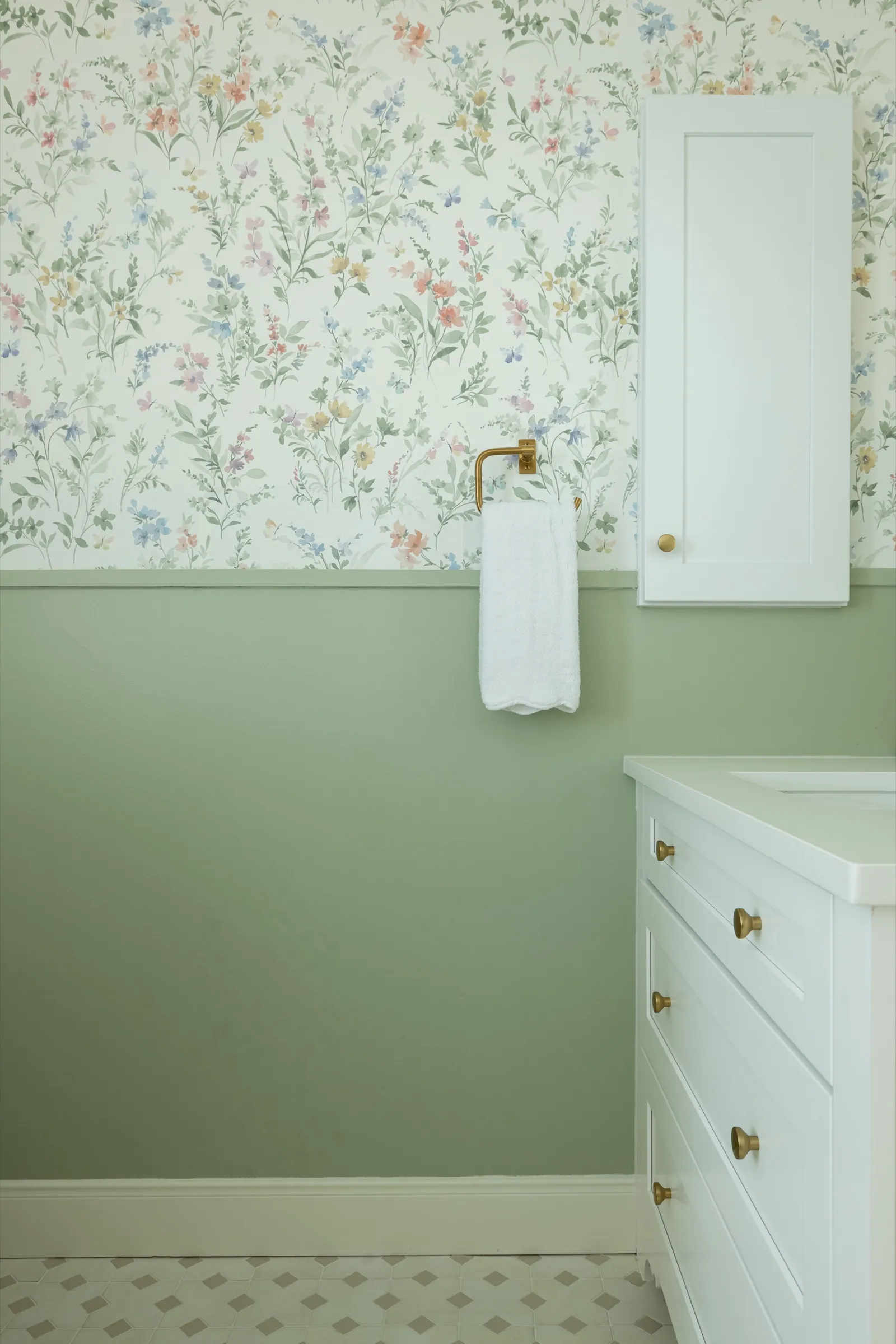 Powder room with wildflower wallpaper above a sage green wainscot, a brass towel ring, a white wall cabinet and a white vanity over a patterned tile floor