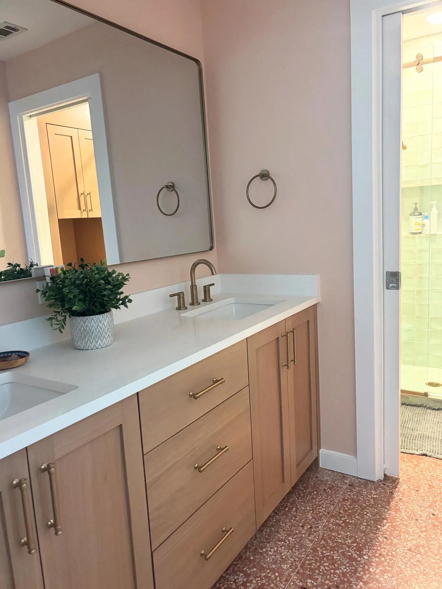White oak double vanity with brass taps and a potted plant against blush pink walls, a pocket door open onto the shower and a terrazzo floor underfoot