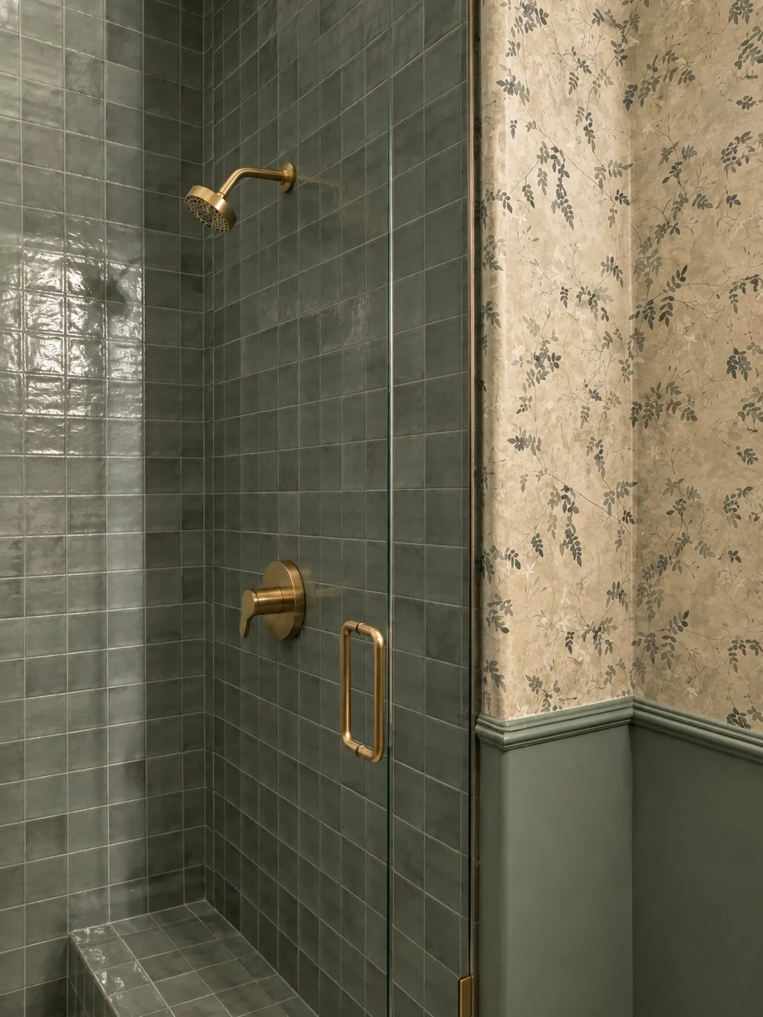 Shower lined in deep green zellige tile with brass fittings, beside the same floral paper and green wainscot