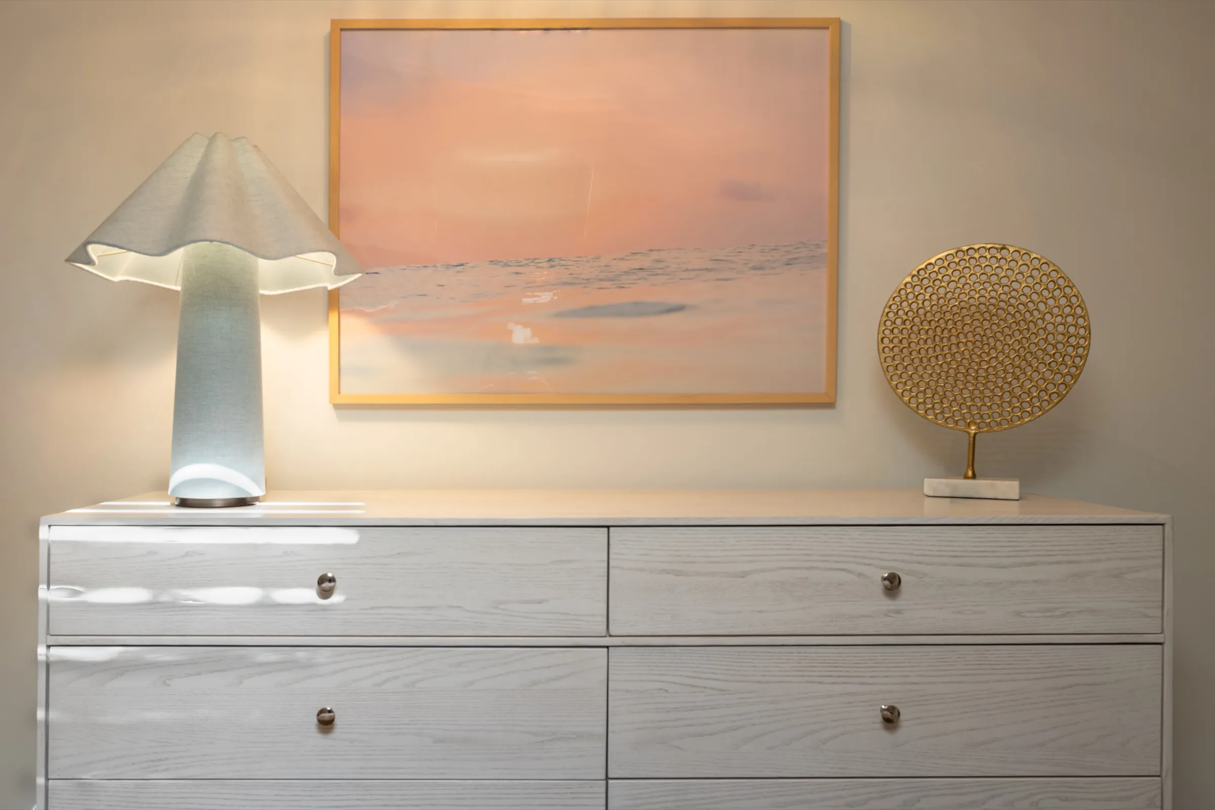 Pale oak dresser with a ruffled ceramic lamp, a framed sunset print and a gilded disc sculpture