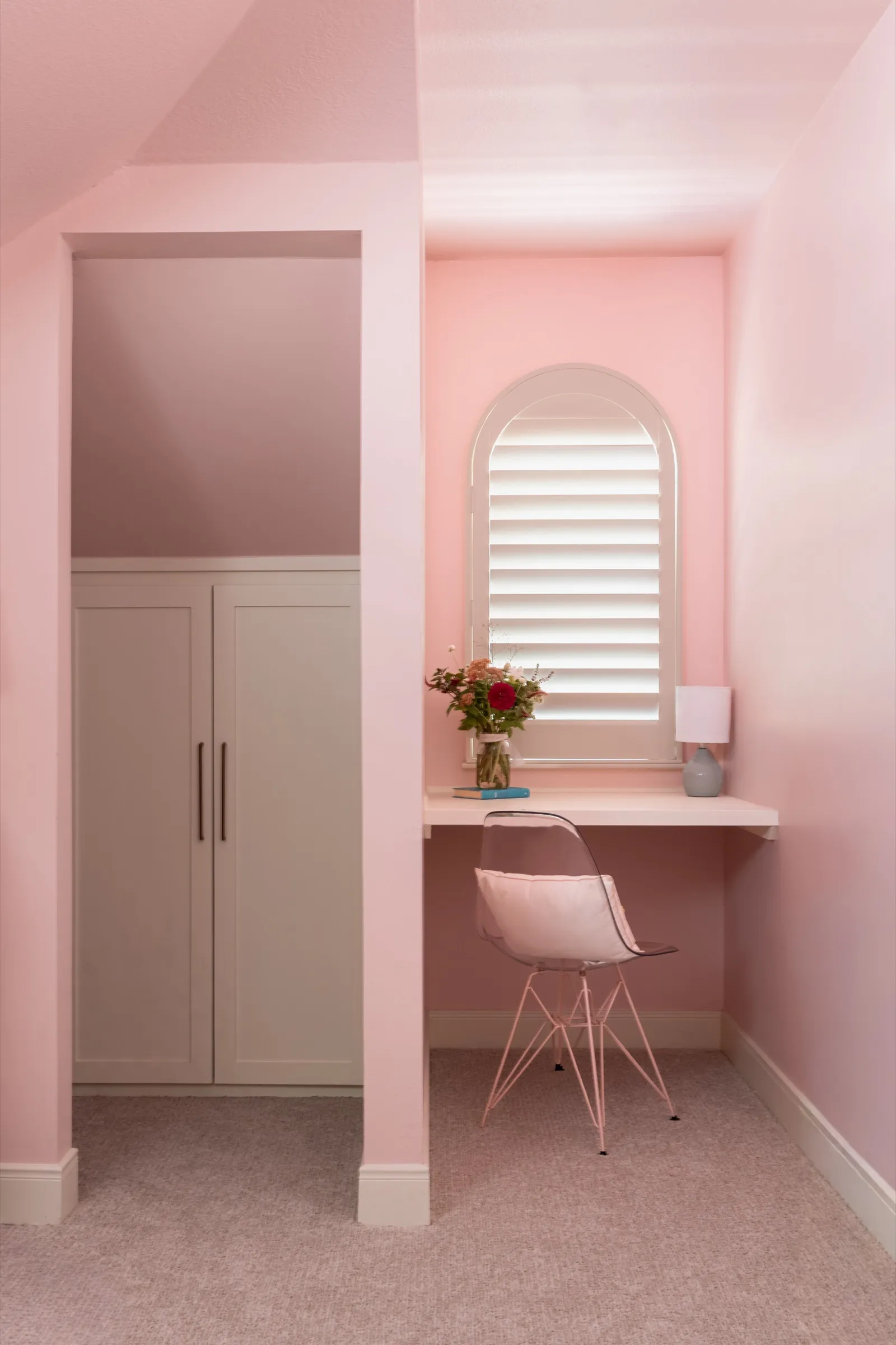 Pink study nook with a built-in desk under an arched shuttered window, a clear chair and a fitted cupboard alongside